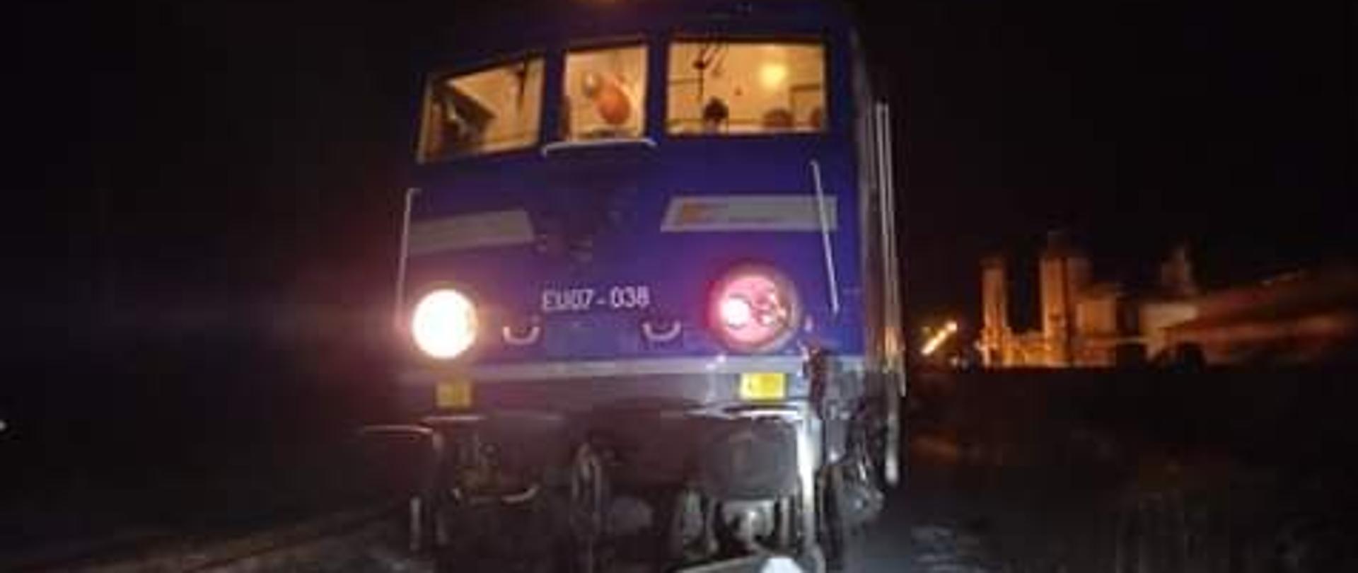 Zdjęcie przedstawia przód lokomotywy pociągu pasażerskiego, lokomotywa koloru niebieskiego z dwoma okrągłymi światłami na przodzie. W tle krajobraz nocny.