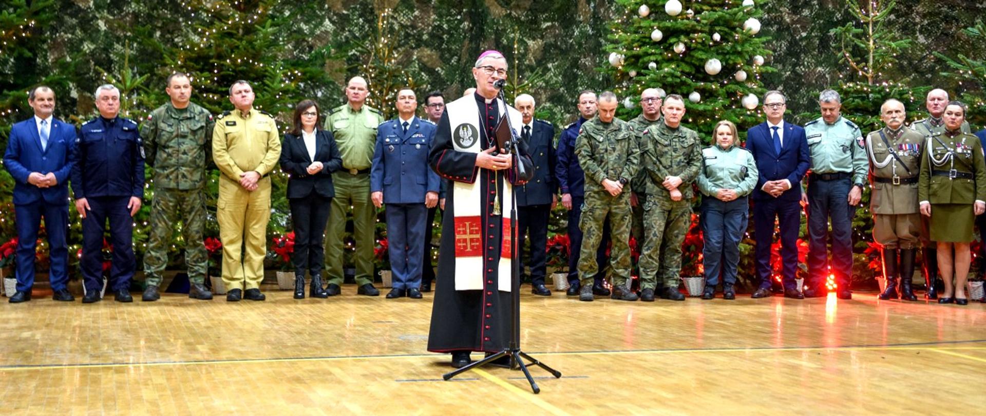 Zdjęcie zrobione w pomieszczeniu. Na zdjęciu uczestnicy Wigilii służb mundurowych. Na pierwszym planie przed mikrofonem stoi biskup rzeszowski. 