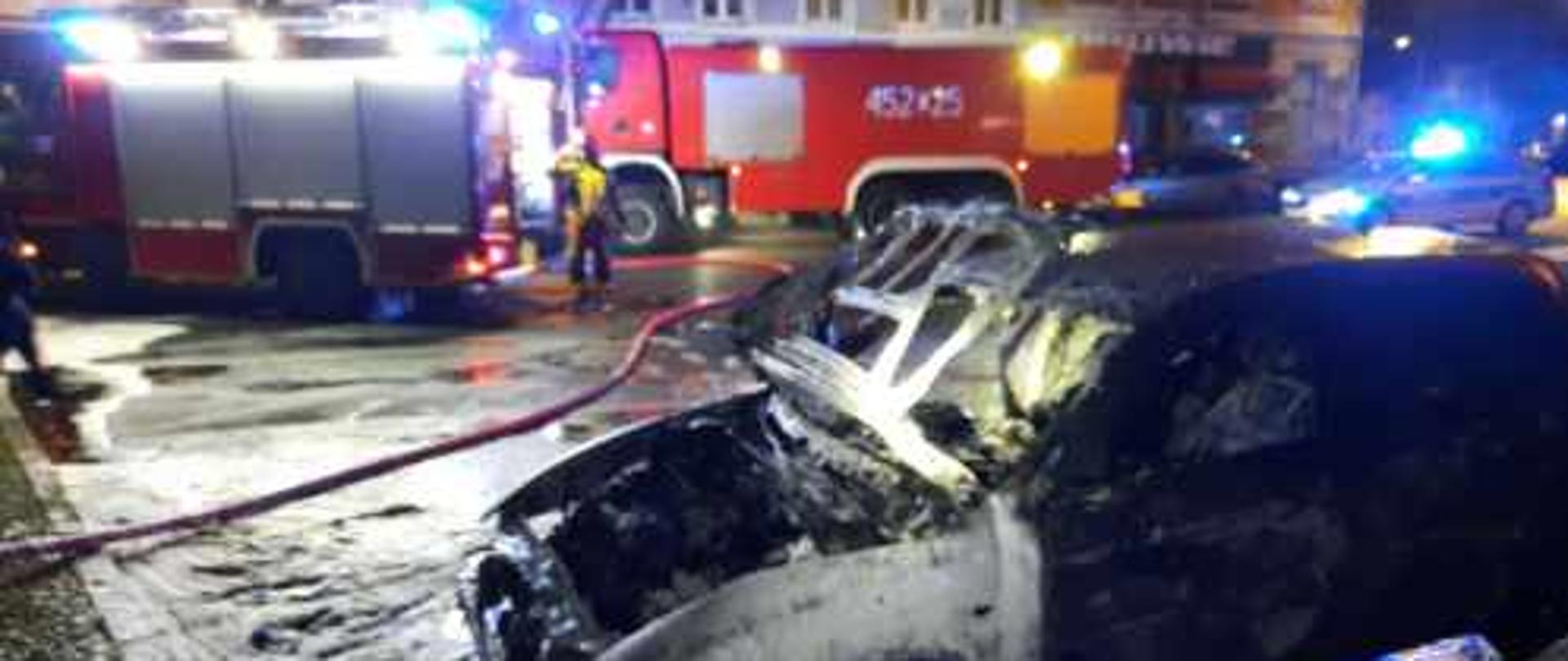 Na parkingu w centrum Lubska powstał pożar od zwarcia instalacji w komorze silnika samochodu. W tle widać strażacki samochód gaśniczy i wrak spalonego samochodu. Pożar powstał w godzinach nocnych.