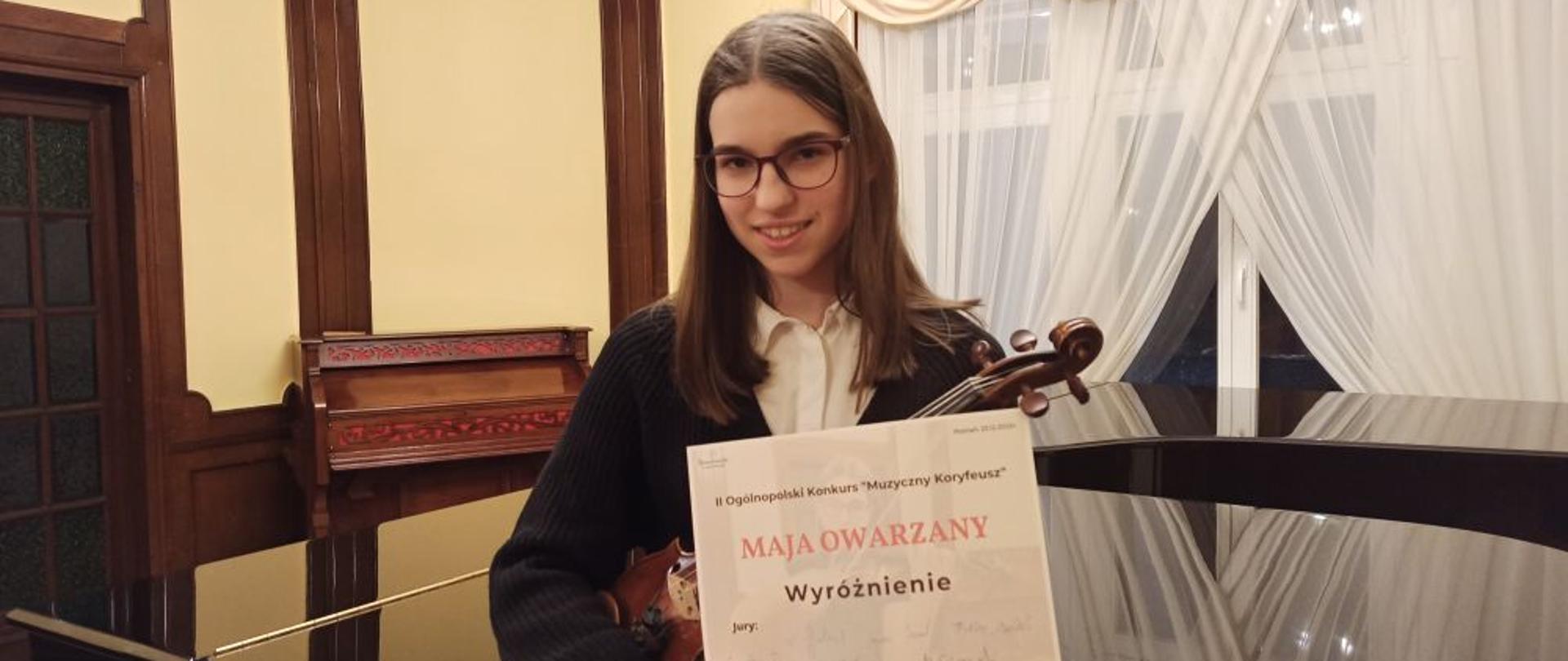 Uczennica ze skrzypcami trzyma w ręku dyplom II Ogólnopolskiego Konkursu "Muzyczny Koryfeusz". Za nią znajduje się fortepian, a z lewej strony stoi fisharmonia.