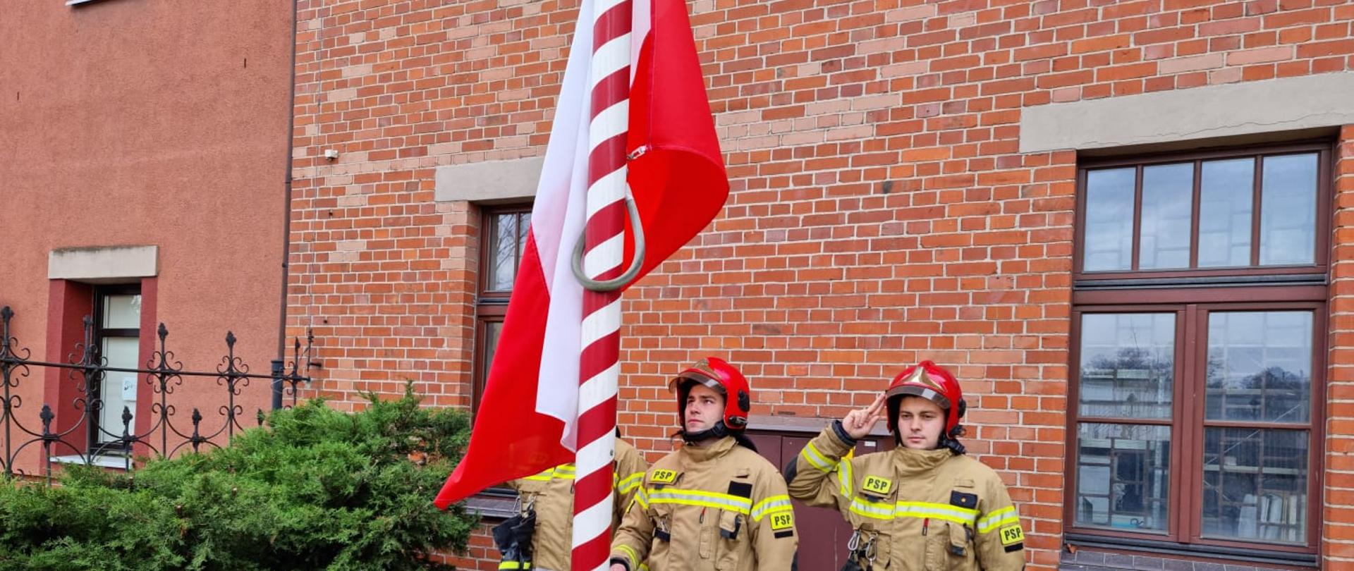 Strażacy w jasnych mundurach i czerwonych hełmach na tle budynku z czerwonej cegły wciągają na masz biało-czerwoną flagę
