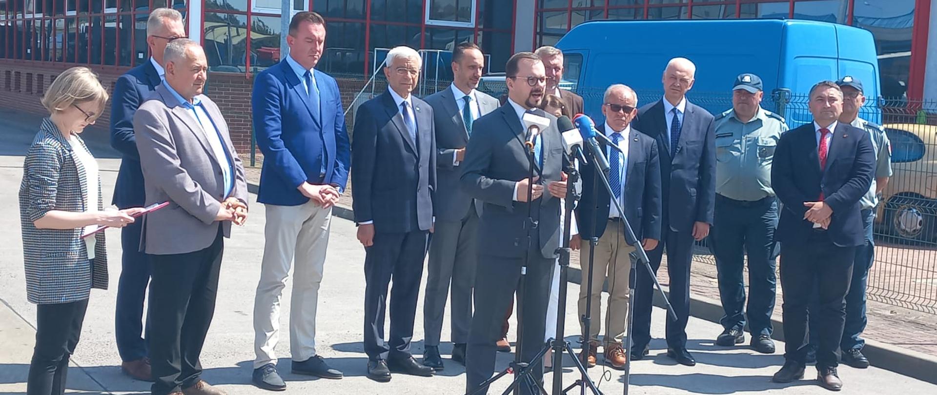 Przemysław Rzodkiewicz zabiera głos przed mikrofonami stojącymi na stojakach, za nim grupa mężczyzn i kobiet stojąca wokół Inspektora