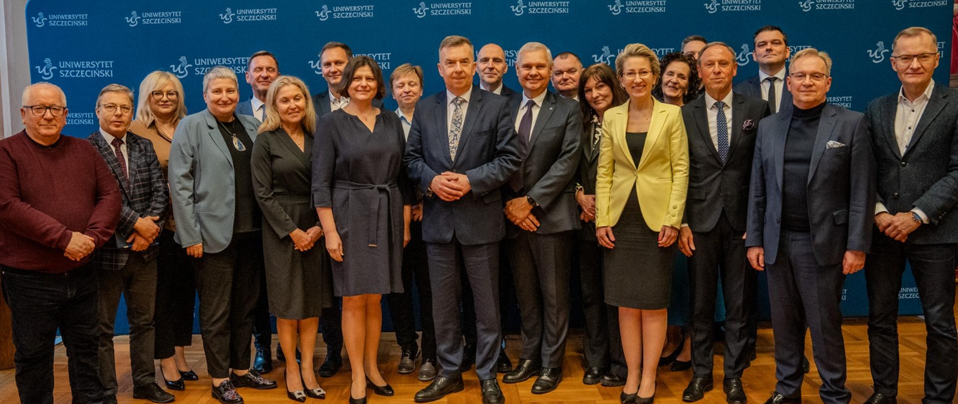 Zdjęcie zbiorowe, kilkanaście osób stoi pod niebieską ścianką z napisem Uniwersytet Szczeciński.