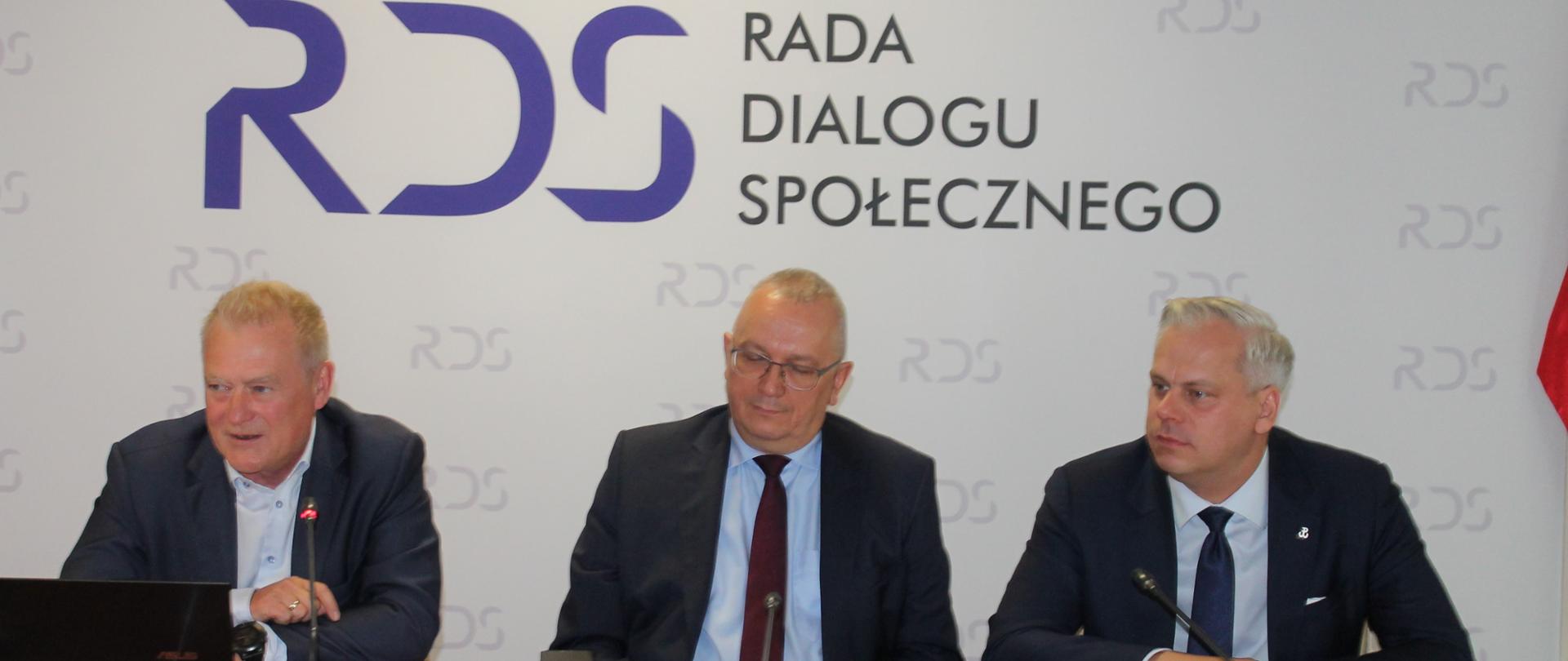 Zdjęcie przedstawia stół prezydialny za którym siedzą: Pan Karol Rabenda – Podsekretarz Stanu w Ministerstwie Aktywów Państwowych, Współprzewodniczący Zespołu ze strony rządowej, Pan Przemysław Kołodziejak – Współprzewodniczący Zespołu ze strony pracodawców oraz Pan Krzysztof Kisielewski – Współprzewodniczący Zespołu ze strony związków zawodowych. W tle znajduje się baner z napisem Rada Dialogu Społecznego