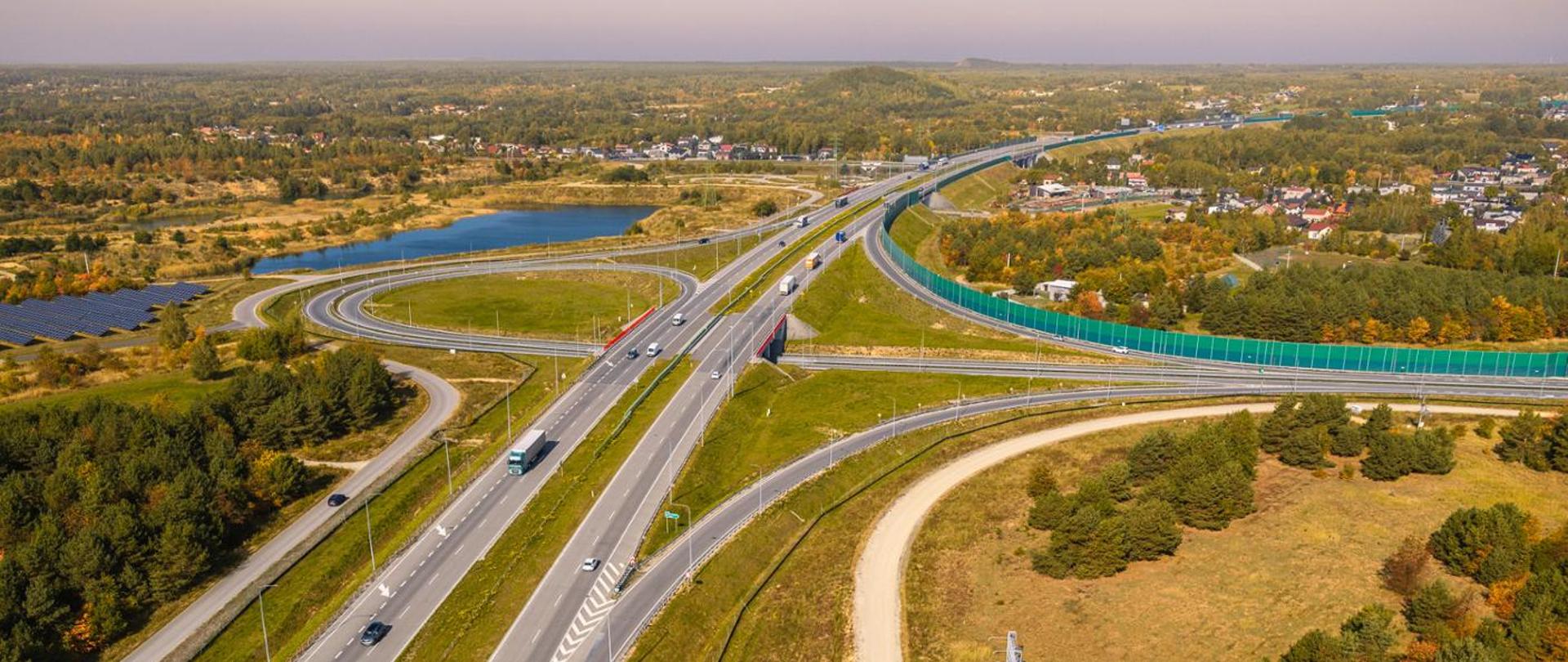 Zdjęcie z lotu ptaka węzła drogowego na autostradzie. Kilkupasmowe jezdnie przecinają się, a wokół nich biegną szerokie, łukowate łącznice. Na drogach widocznych jest kilka samochodów osobowych i ciężarowych. W otoczeniu znajdują się tereny zielone, zadrzewienia oraz niewielki zbiornik wodny. Po lewej stronie widoczna jest instalacja fotowoltaiczna, a po prawej ekran akustyczny oddzielający drogę od zabudowy mieszkaniowej. W tle rozciąga się rozległy, częściowo zalesiony krajobraz z rozproszoną zabudową. Zdjęcie wykonano w słoneczny dzień z dużej wysokości.