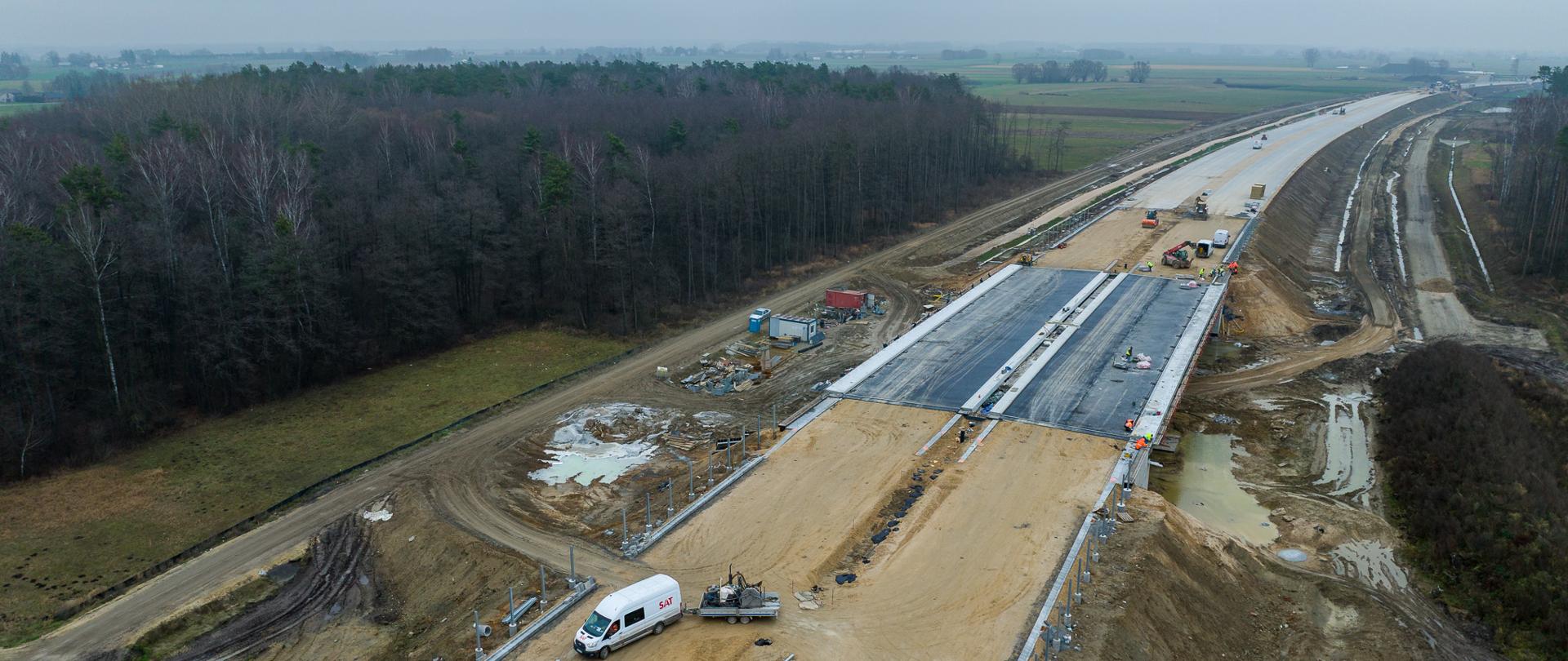 Budowa odcinka drogi ekspresowej – widok z góry na plac budowy z ułożonymi warstwami podbudowy, maszynami drogowymi i pojazdami serwisowymi, otoczony terenami leśnymi i polami.