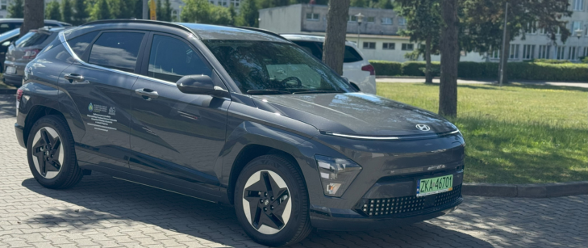 Zdjęcie przedstawia samochód Hyundai Kona w kolorze szarym na tle pasa zieleni, drzew i kilku samochodów.