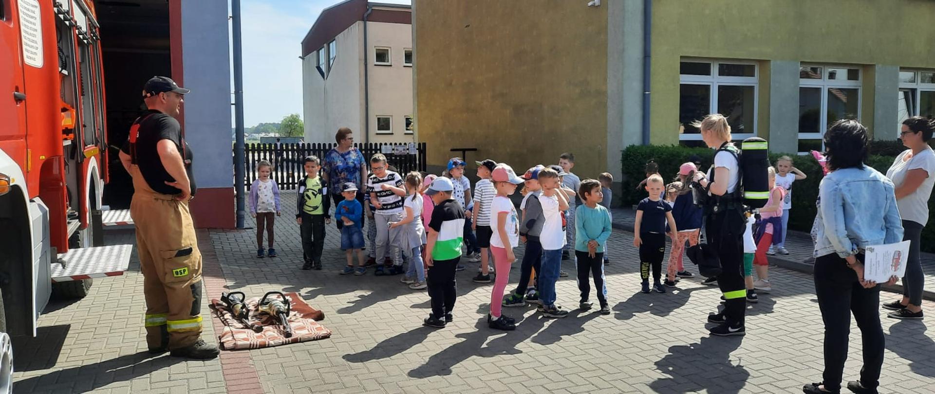 Na zdjęciu znajdują się dzieci, opiekunowie oraz strażacy ubrani w ubrania strażackie na tle budynków i pojazdu strażackiego