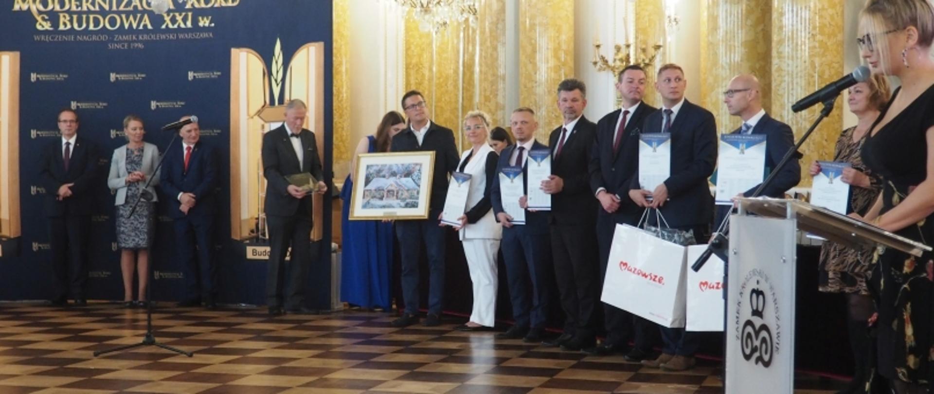 Grupa osób na scenie podczas gali; w tle baner z napisem „Modernizacja Roku & Budowa XXI w.”, niektórzy trzymają dyplomy i torby z napisem „Mazowsze”.