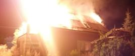 Zdjęcie wykonane w nocy. Praktycznie cały kadr zajmuje ogromny płomień, który opanował mały budynek mieszkalny. Ogień jest potężny, żółty, bardzo wysoki.