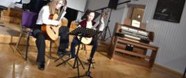 Na zdjęciu widnieje duet gitarowy złożony z uczennic szkoły. Zdjęcie wykonane jest w auli PSM. W tle fortepian oraz organy. Kolorystyka zdjęcia jest biało-brązowa.