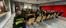 Strażak (funkcjonariusz KP PSP Węgrów) w ubraniu służbowym przemawia do siedzących strażaków w ubraniu koszarowych. Spotkanie jest w sali konferencyjnej siedziby KP PSP w Węgrowie. W tle po lewej stronie widać dwie flagi Polski oraz flagę UE.