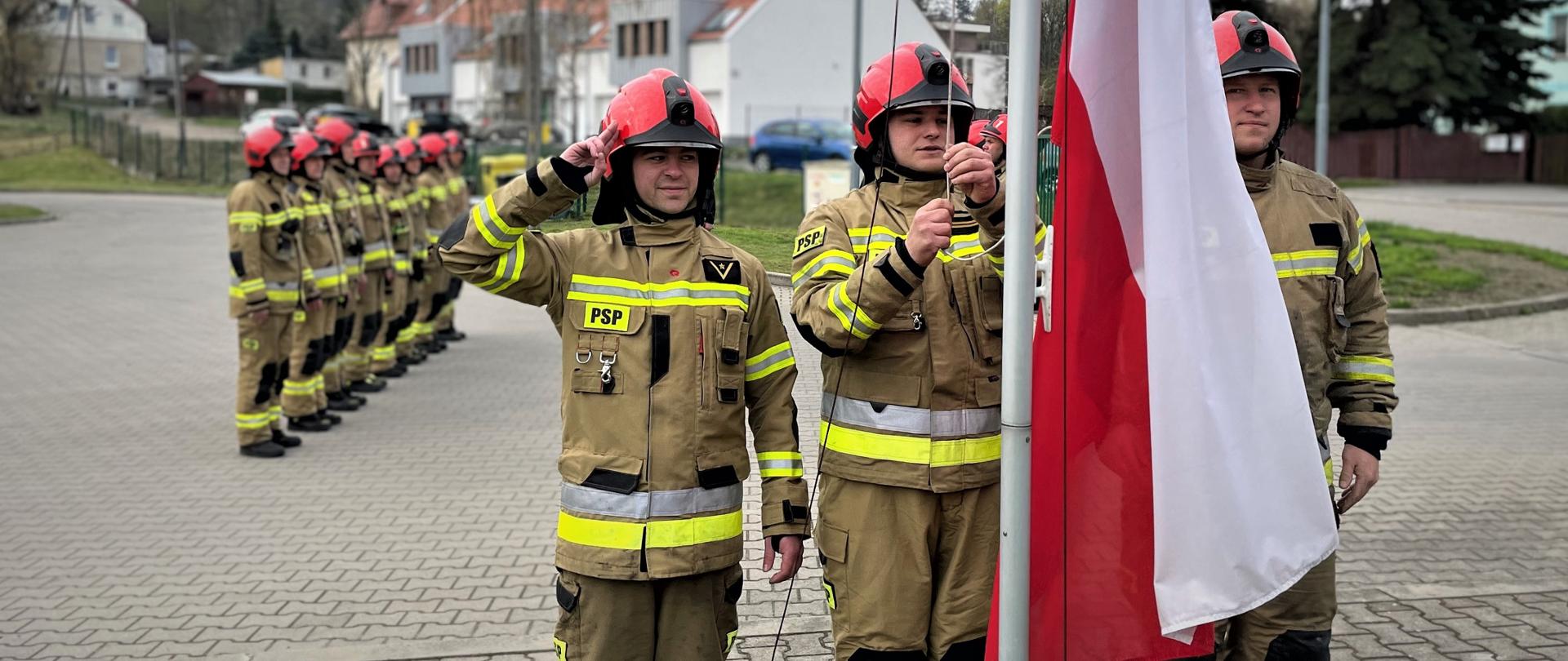 Na zdjęciu widać strażaków przed masztem na którym zawieszają flagę państwową. Za strażakami stoją dwa rzędy zmiany zdającej i przyjmującej służbę. Strażacy w umundurowaniu bojowym. 