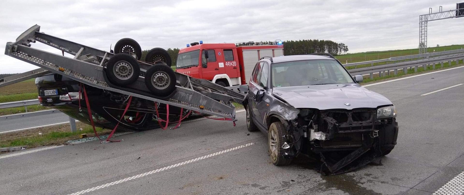 Na zdjęciu widać kolizję drogową na drodze. Jedne auto leży na dachu, przyciśnięte przyczepą, drugie auto ma uszkodzony przód. W tle pojazd pożarniczy.