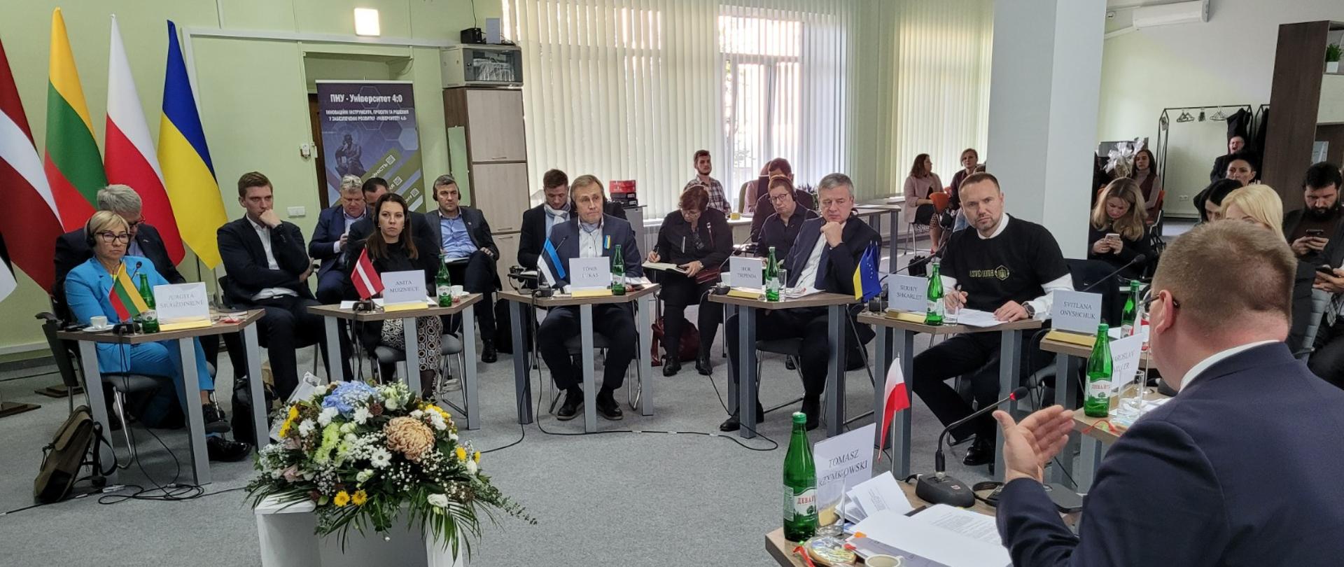 Grupa osób, które w półkolu siedzą przy małych stolikach. Na stolikach kartki z nazwiskami, dokumenty, napoje. Na pierwszm planie stolik z kwiatami. W lewym rogu flagi. 