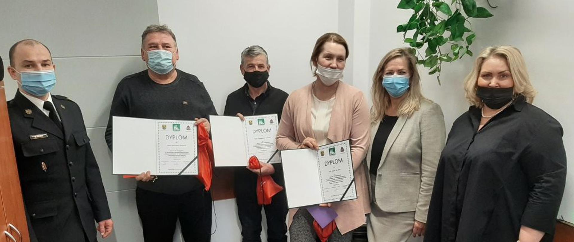 W pomieszczeniu na tle białej ściany stoją 3 kobiety i trzech mężczyzn w maseczkach. Trzy osoby po środku trzyma przed sobą dyplomy.