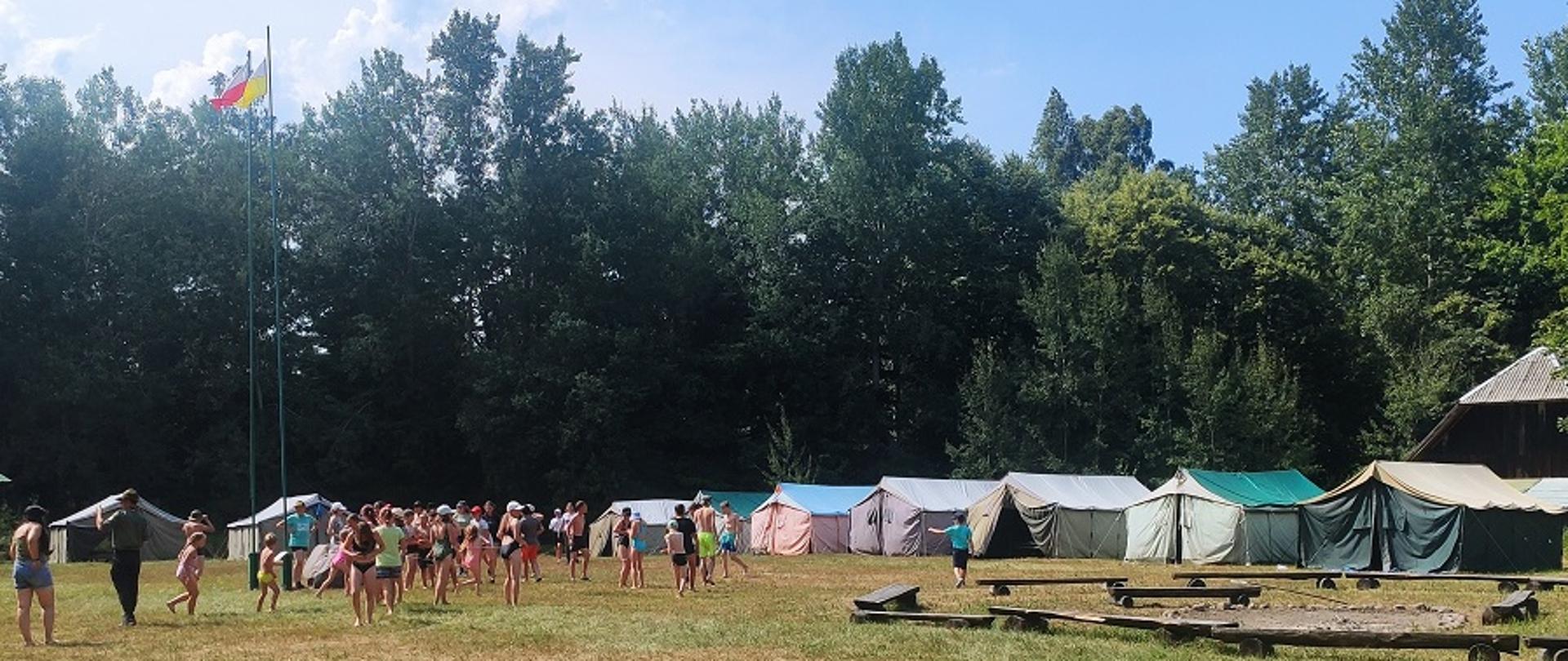 Obozowisko harcerskie. Z lewej maszty z flagami m. in. Polski a przy nich grupa dzieci. W tle namioty harcerskie.