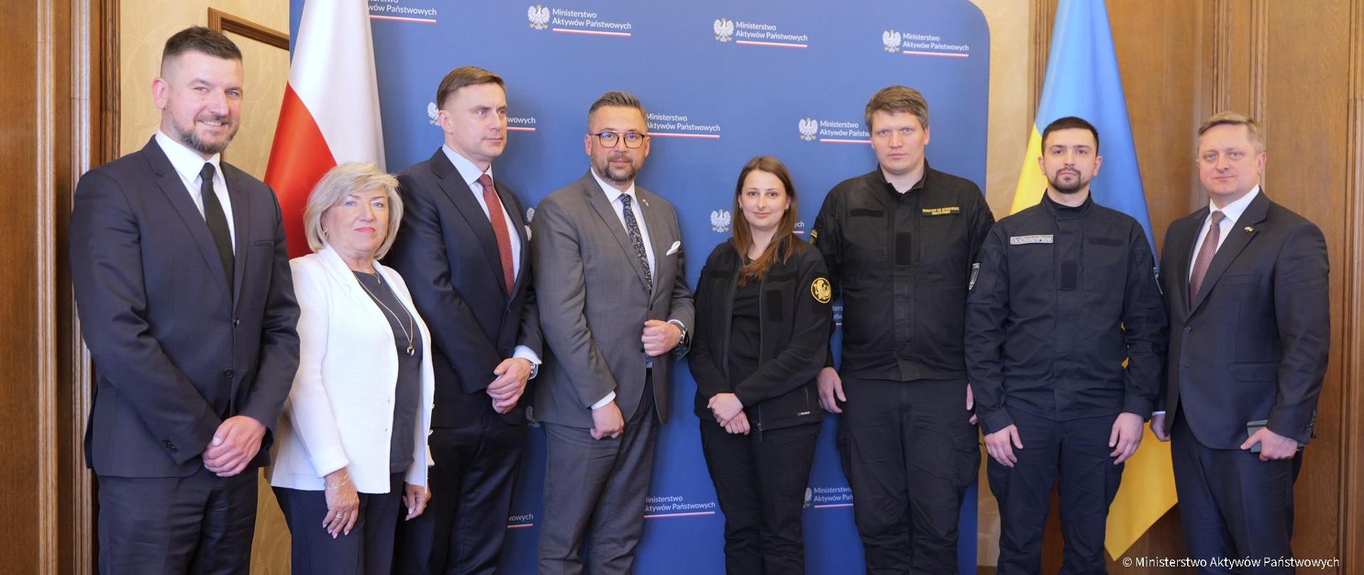 Uczestnicy spotkania stoją na tle flag Ukrainy i Polski. Wiceminister Marcin Kulasek w środku.