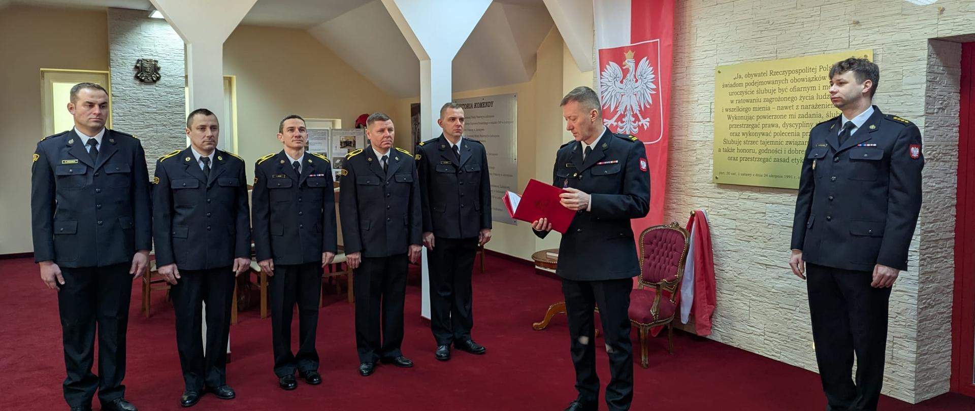 Komendant Powiatowy PSP w Jędrzejowie odczytuje decyzje o powołaniu na wyższe stanowiska służbowe. Po jego prawej stronie 5 awansowanych funkcjonariuszy w umundurowaniu wyjściowym. 