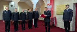 Komendant Powiatowy PSP w Jędrzejowie odczytuje decyzje o powołaniu na wyższe stanowiska służbowe. Po jego prawej stronie 5 awansowanych funkcjonariuszy w umundurowaniu wyjściowym. 