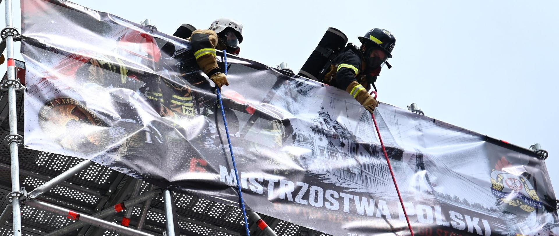 Dwóch strażaków w ubraniach specjalnych pokonuje przeszkodę na wysokości podczas zawodów strażackich. W tle widoczny baner Mistrzostw Polski.