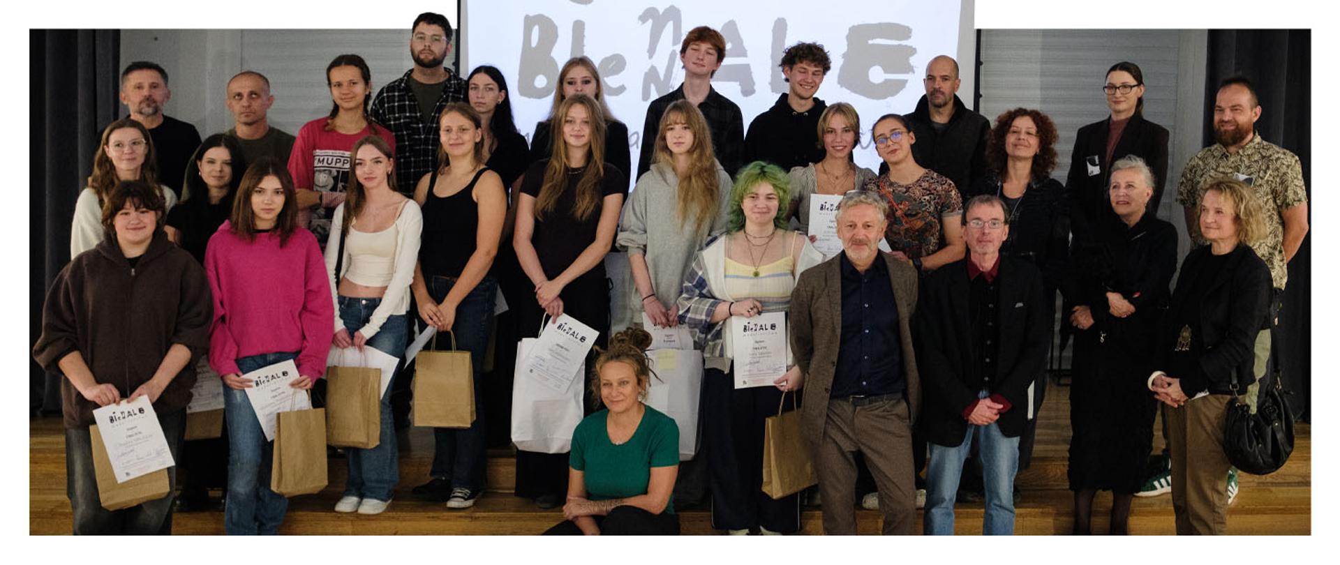 Pamiątkowe zdjęcie grupowe wszystkich laureatów, jurorów i organizatorów VI Biennale Medalierstwa. Uczestnicy stoją na schodach sceny, dumnie prezentując dyplomy i nagrody. 