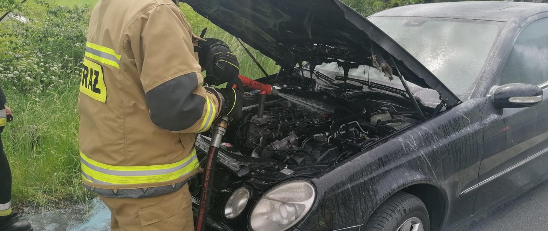 Widoczne miejsce zdarzenia uszkodzony samochód oraz strażacy prowadzący działania gaśnicze. 