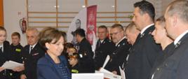 Wręczenie promes. Sala sportowa w Szkaradowie. Pani minister Marlena Maląg wręcza promesy strażakom OSP. Kobieta ma wyciągniętą prawą dłoń do złożenia gratulacji. W lewej ręce trzyma białą teczkę. W tle inni uczestnicy uroczystości. 
