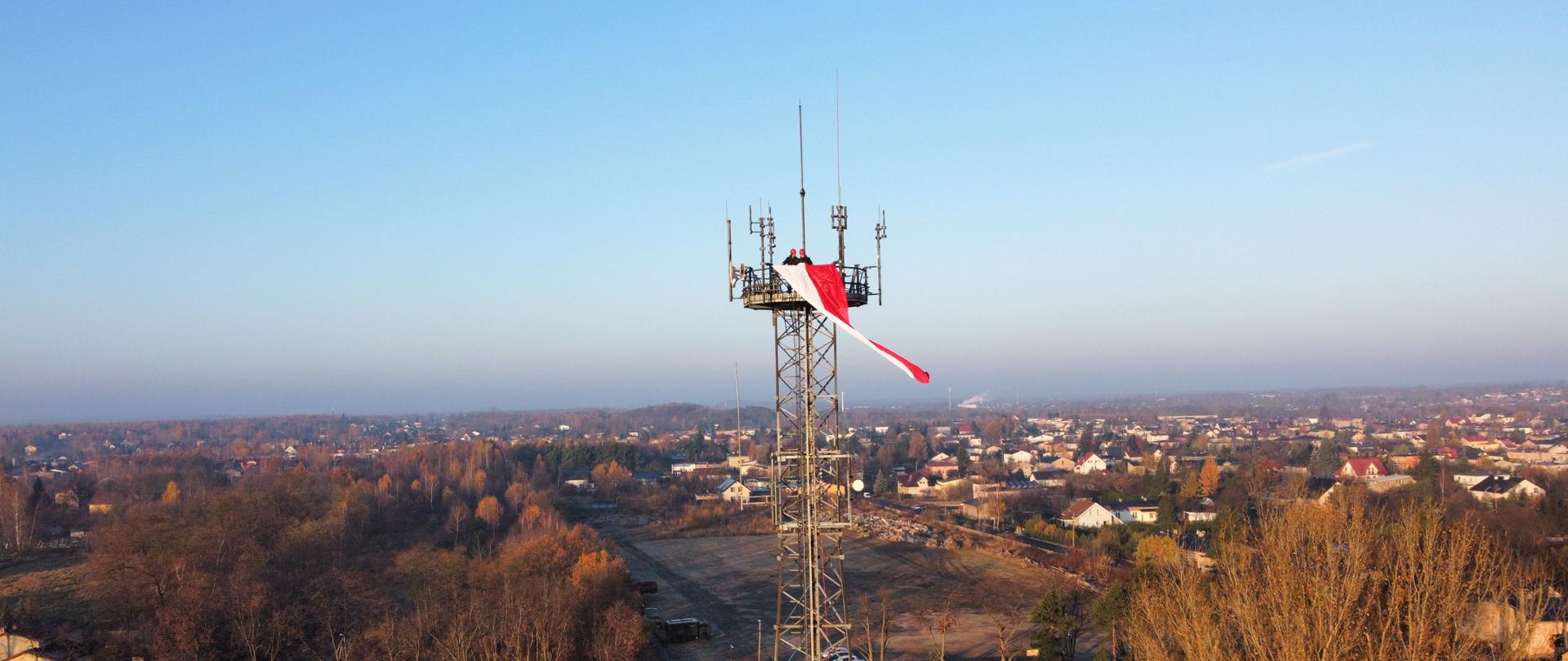 Dwóch strażaków zawiesza flagę narodową na maszcie antenowym kratownicowym, w tle panorama dzielnicy Stradom w Częstochowie