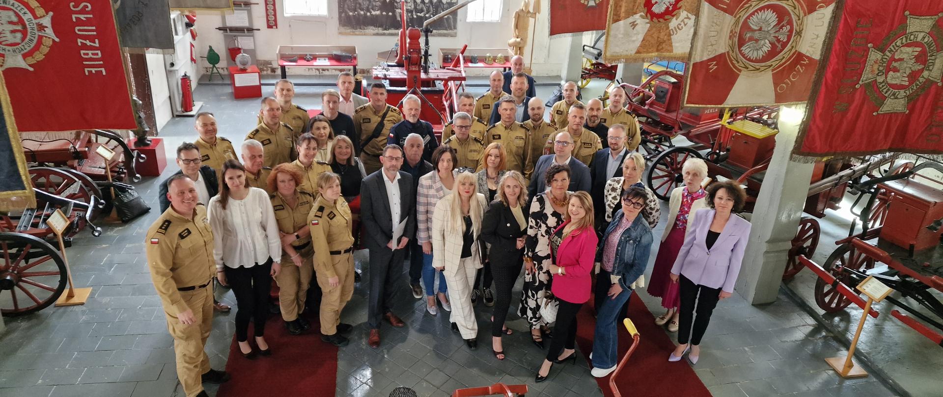Zdjecie grupowe w muzeum pożarnictwa, grupa strażaków i nauczycieli pomiędzy starymi wozami straży pożarnej, nad nimi wiszą sztandary