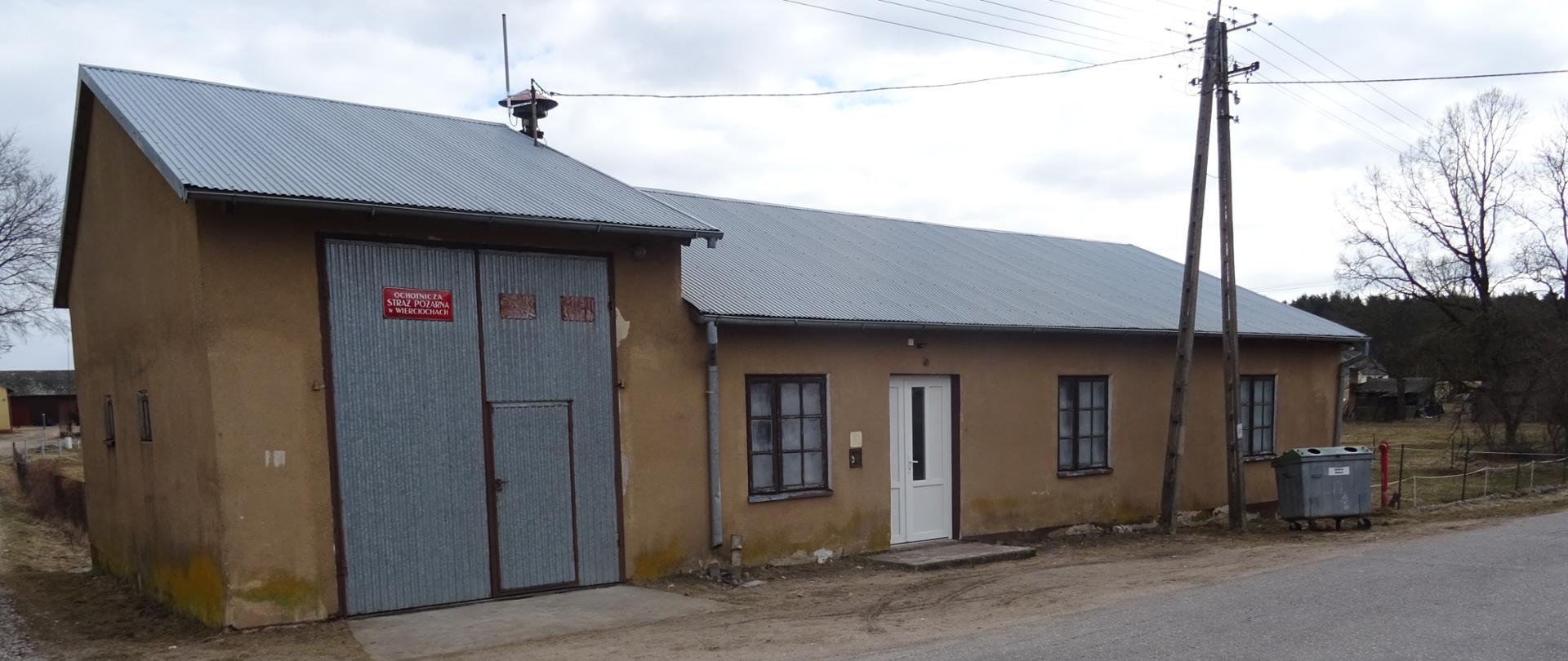 Na zdjęciu znajduje się budynek remizy OSP Wierciochy budynek parterowy o szarych ścianach i spadzistym dachu na którym umieszczona jest syrena
