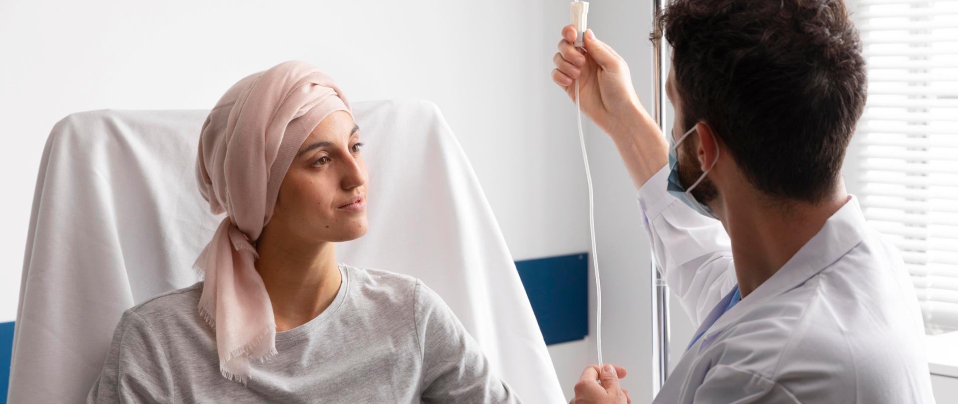 Zdjęcie przedstawia pacjentkę chorą na nowotwór, która leży na łóżku. na głowie na turban. Przy niej siedzi lekarz, który reguluje dopływ dożylny leków. 