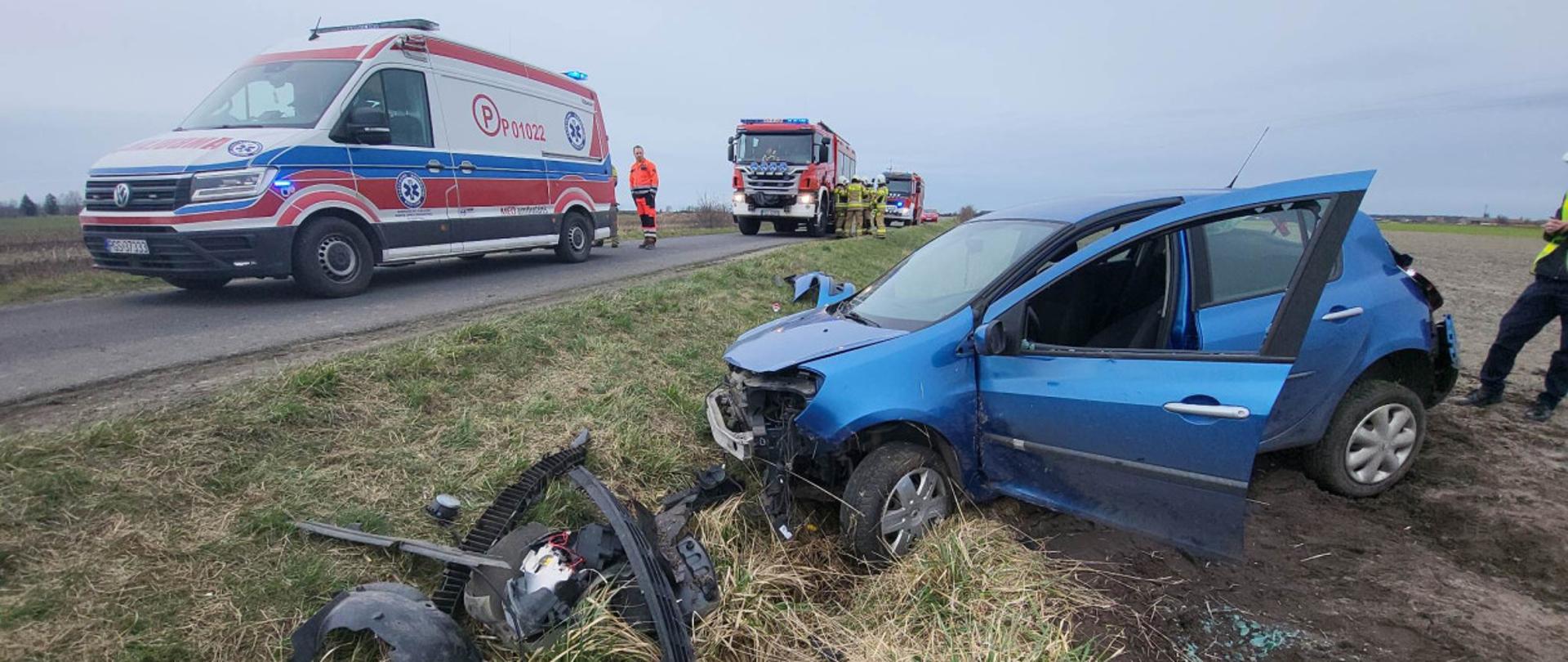 Zdjęcie przedstawia samochód osobowy po kolizji. Znajduje on się na poboczu (częściowo na polu). Samochód jest zniszczony. W tle znajduje się karetka pogotowia oraz 2 wozy straży pożarnej. Przy karetce stoi ratownik medyczny, a przy wozach strażackich druhowie OSP.