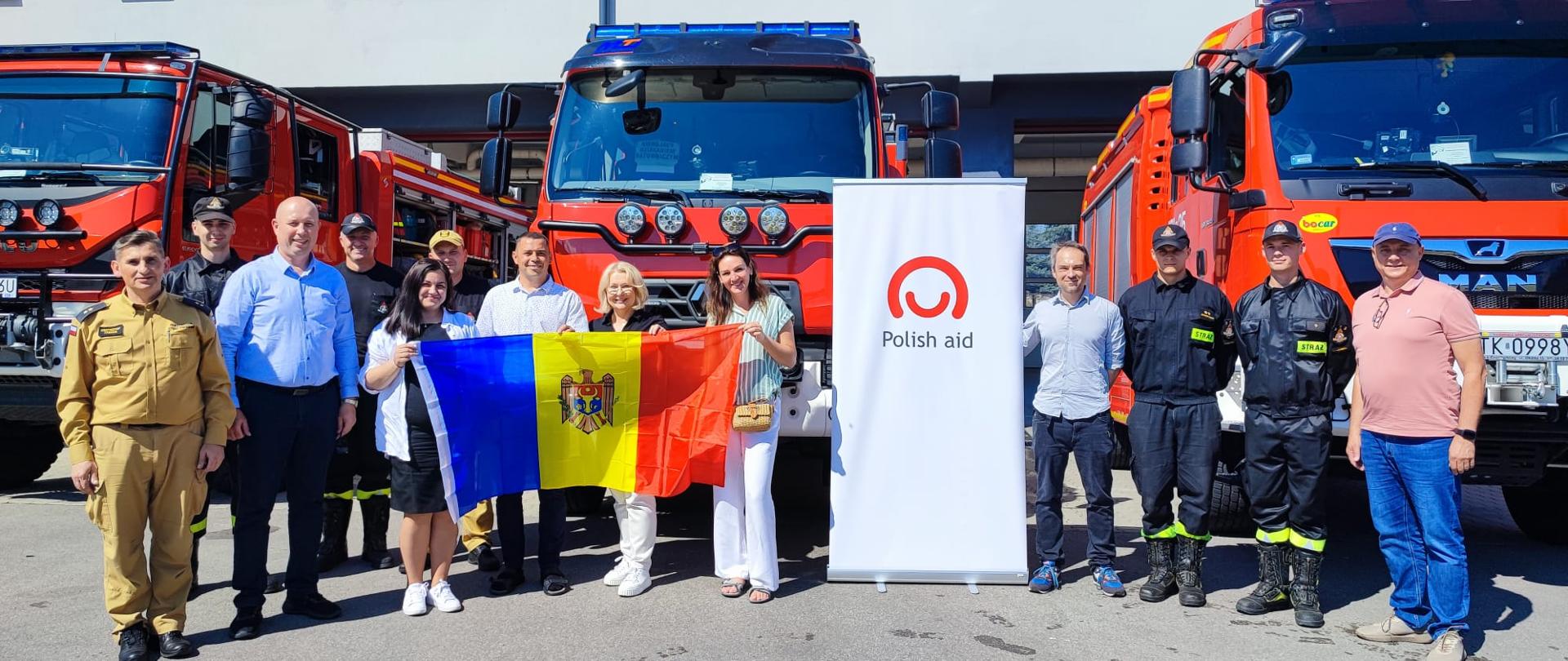 Grupa kobiet i mężczyzn, część w strojach strażackich, pozuje na tle wozów strażackich, z flagą Mołdawii i banerem z logo Polskiej pomocy