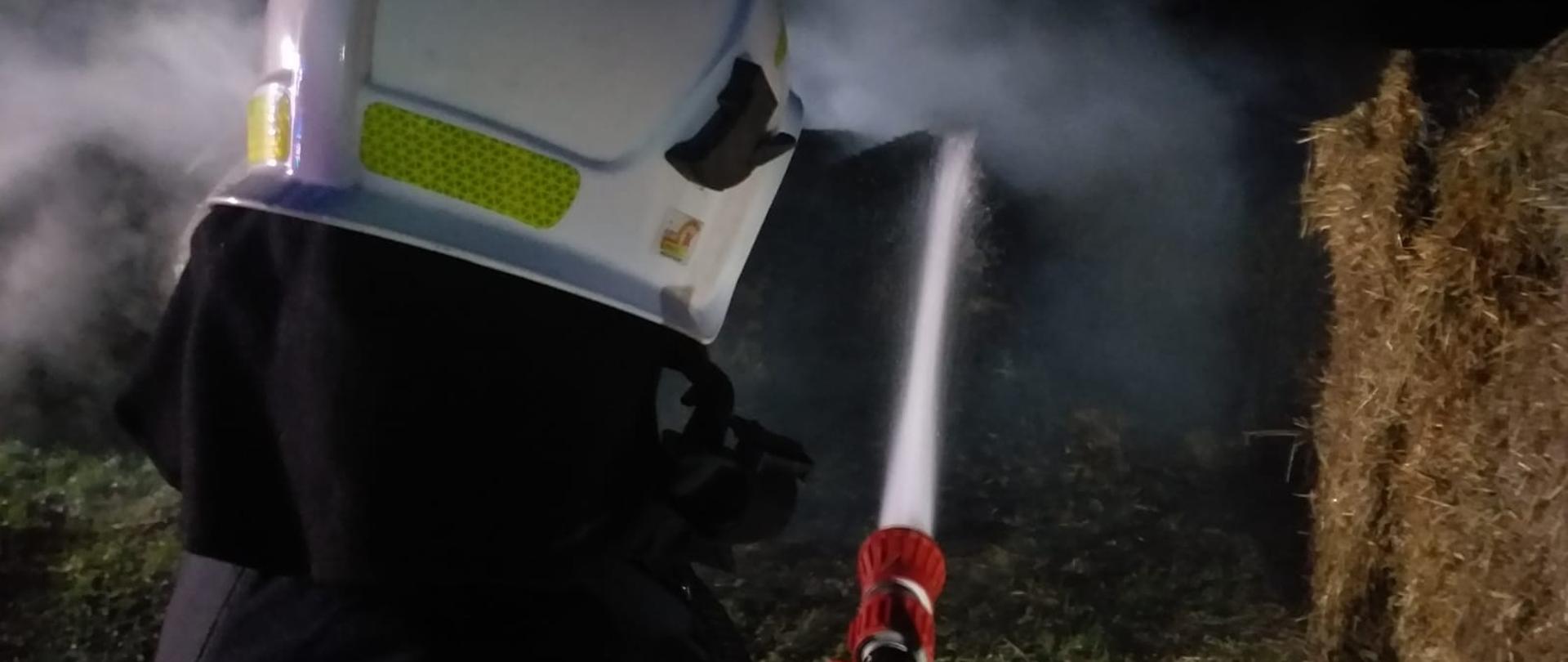 Zdjęcie przedstawia strażaka odwróconego plecami do fotografa. Strażak w umundurowaniu bojowym z białym hełmem na głowie trzyma w rękach prądownicę wodną, z której wydobywa się woda. W tle widać baloty słomy, z których unosi się niewielkie zadymienie. Zdjęcie zrobione w porze nocnej.
