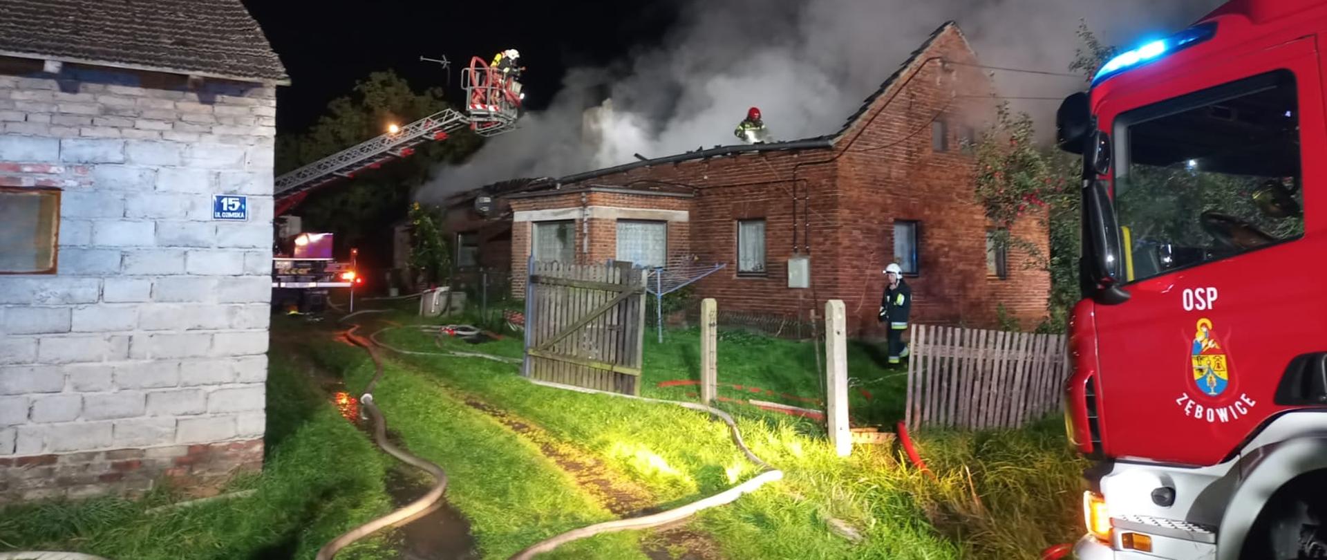 Pożar dachu budynku mieszkalnego, parterowego z czerwonej cegły. Dach budynku zarwany, z budynku wydobywa się biały dym. Obok budynku stoją samochody pożarnicze. 