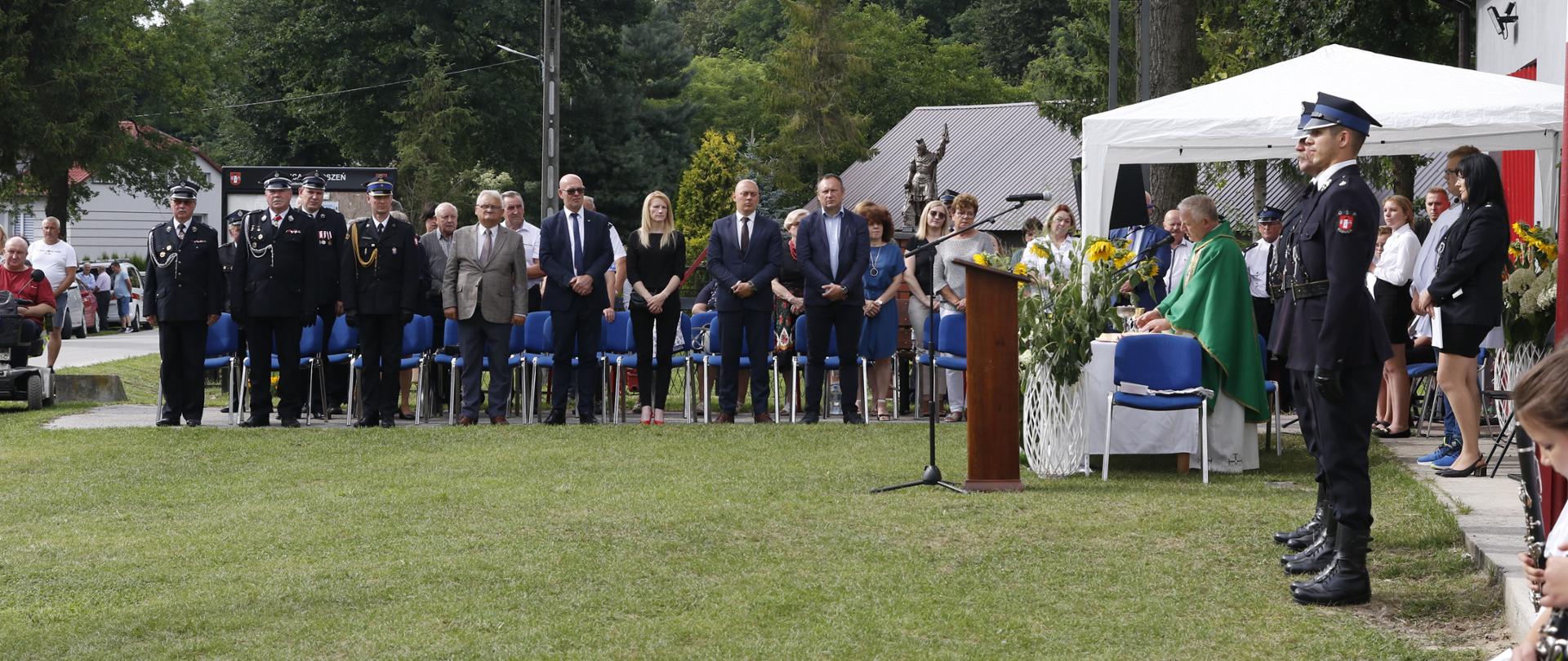 Zdjęcie przedstawia zaproszonych gości honorowych. Po prawej stronie ołtarz polowy, przy którym ksiądz odprawia mszę świętą w intencji strażaków i ich rodzin. 