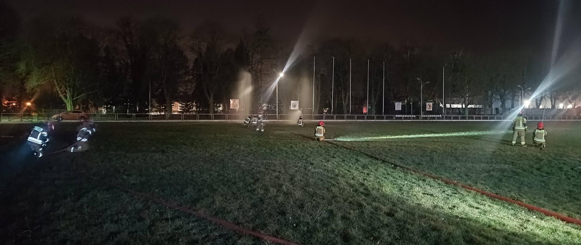 Nocne manewry taktyczne, na murawie stadionu ćwiczą zastępy strażaków, przy każdym zastępie widoczna doprowadzona linia wodna