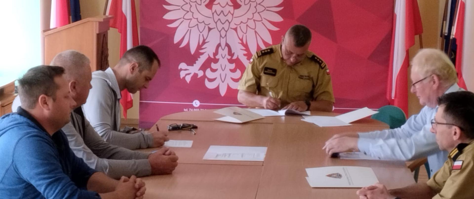 Sala konferencyjna w Komendzie Powiatowej Państwowej Straży Pożarnej w Kłodzku. Przy stole konferencyjnym Komendant wraz z wykonawcą podpisują umowę na wykonanie zadania.