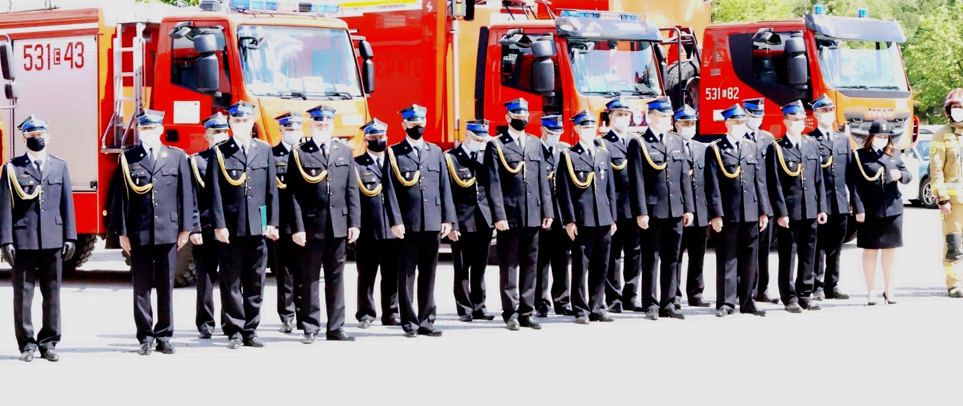 Na zdjęciu widać strażaków stojących w szyku podczas akademii z okazji dnia strażaka. W tle wozy strażackie