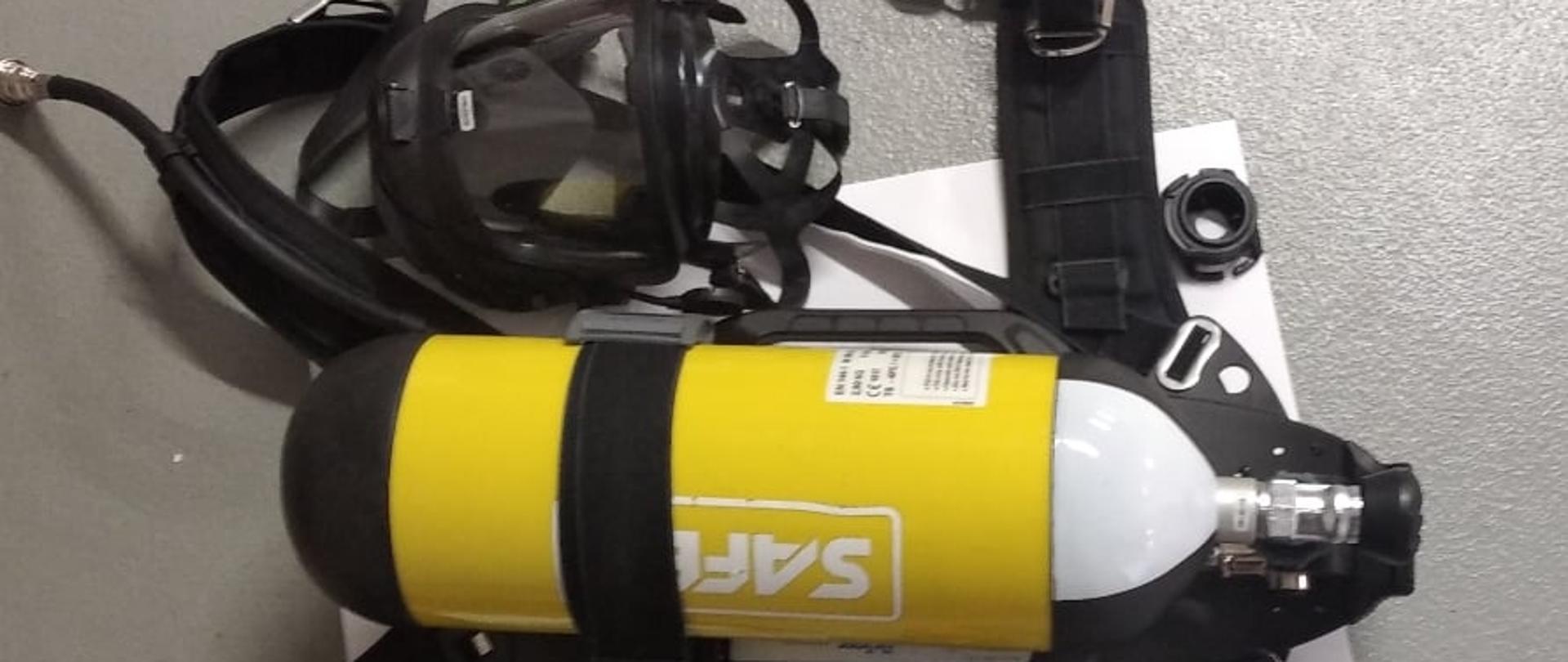 Aparat powietrzny składający się z zółtej butli ze stelażem oraz maski pełnotwarzowej leży na szarej posadzce w garażu.