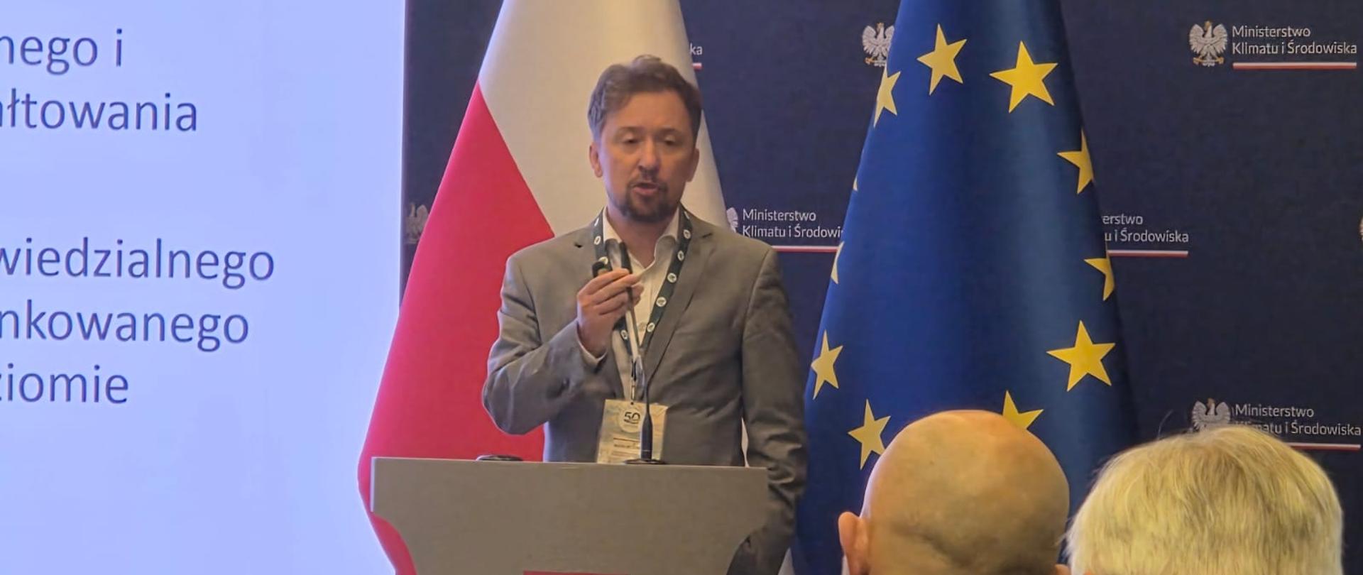 Na zdjęciu Piotr Otawski - Generalny Dyrektor Ochrony Środowiska, który przemawia, stojąc za mównicą z godłem Polski. Za jego plecami znajdują się dwie duże flagi: polska oraz Unii Europejskiej, a także tło z powtarzającym się logo Ministerstwa Klimatu i Środowiska. Po lewej stronie wyświetlany jest fragment prezentacji. W pierwszym planie widoczne są tyły głów dwóch siedzących mężczyzn, którzy uczestniczą w tym spotkaniu.