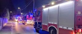 Zdjęcia w porze nocnej. Widać pojazdy straży pożarnej przed posesją gdzie doszło do pożaru. Sygnalizacja ostrzegawcza włączona. Droga publiczna. Lampy oświetlenia ulicznego włączone.