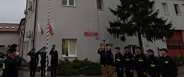 Strażacy stoją na placu przed jednostką w dwóch szeregach na przeciw siebie. Przy maszcie stoją strażacy i oddają hołd. Na maszcie wisi flaga państwowa. 