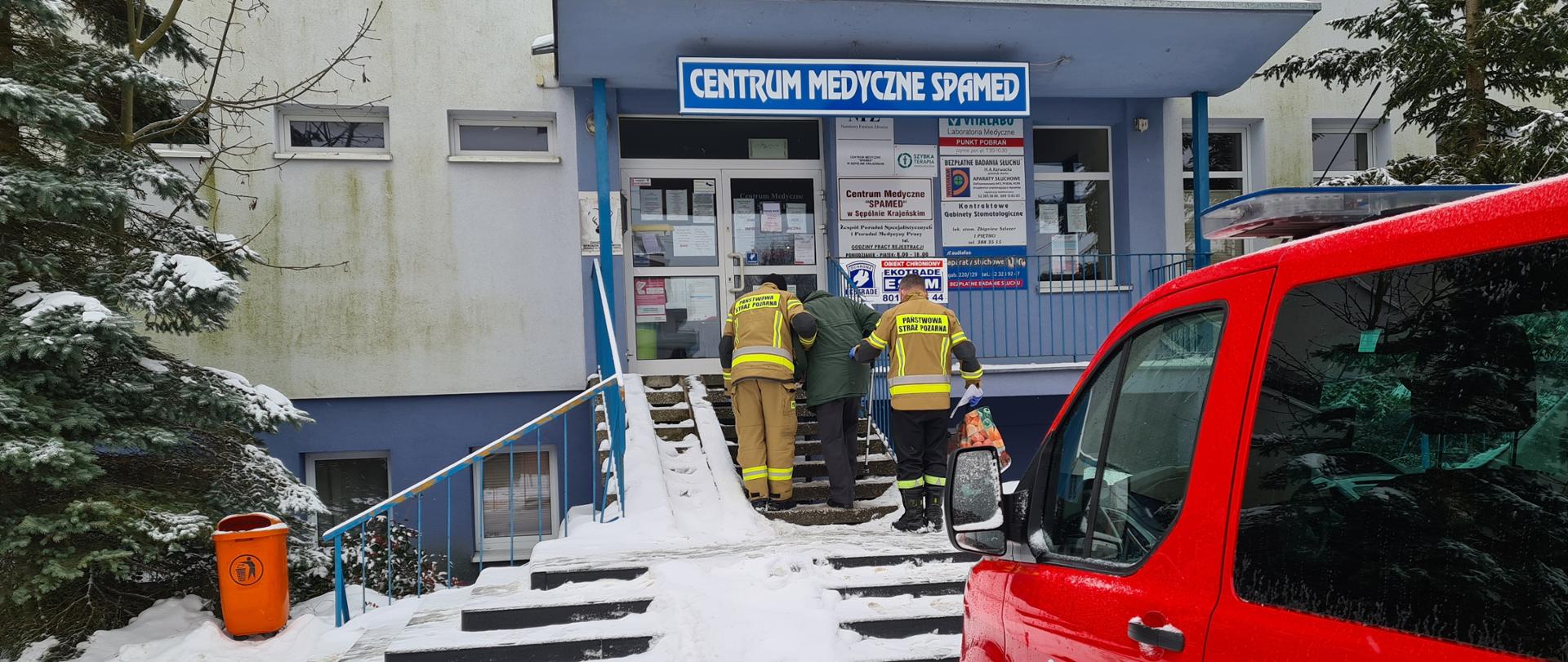 Zdjęcie wykonano za dnia zimową porą przed przychodnią Spamed w Sępólnie krajeńskim. Widzimy na nim na pierwszym planie po prawej samochód straży pożarnej typu bus koloru czerwonego. Na dalszym planie dwóch strażaków trzymając pod pachą starszą osobę, pomagają jej wejść po schodach do punktu szczepień