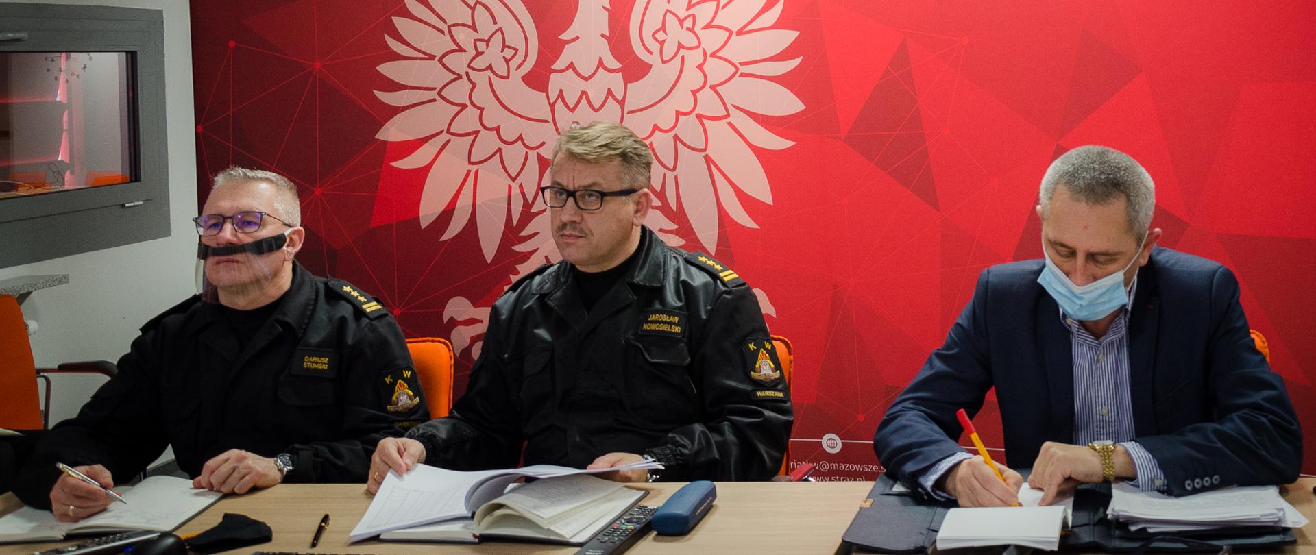 Zdjęcie przedstawia trzy osoby, w tym dwóch strażaków oraz jedną osobę cywilną podczas wideokonferencji. W tle ścianka koloru czerwonego oraz duże godło RP w kolorze białym.