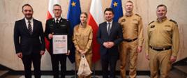 Sześć osób pozuje do pamiątkowego zdjęcia na tle flag Polski i Unii Europejskiej podczas wręczenia dyplomu i upominku strażakowi w galowym mundurze oraz młodej dziewczynie w mundurze beżowym.