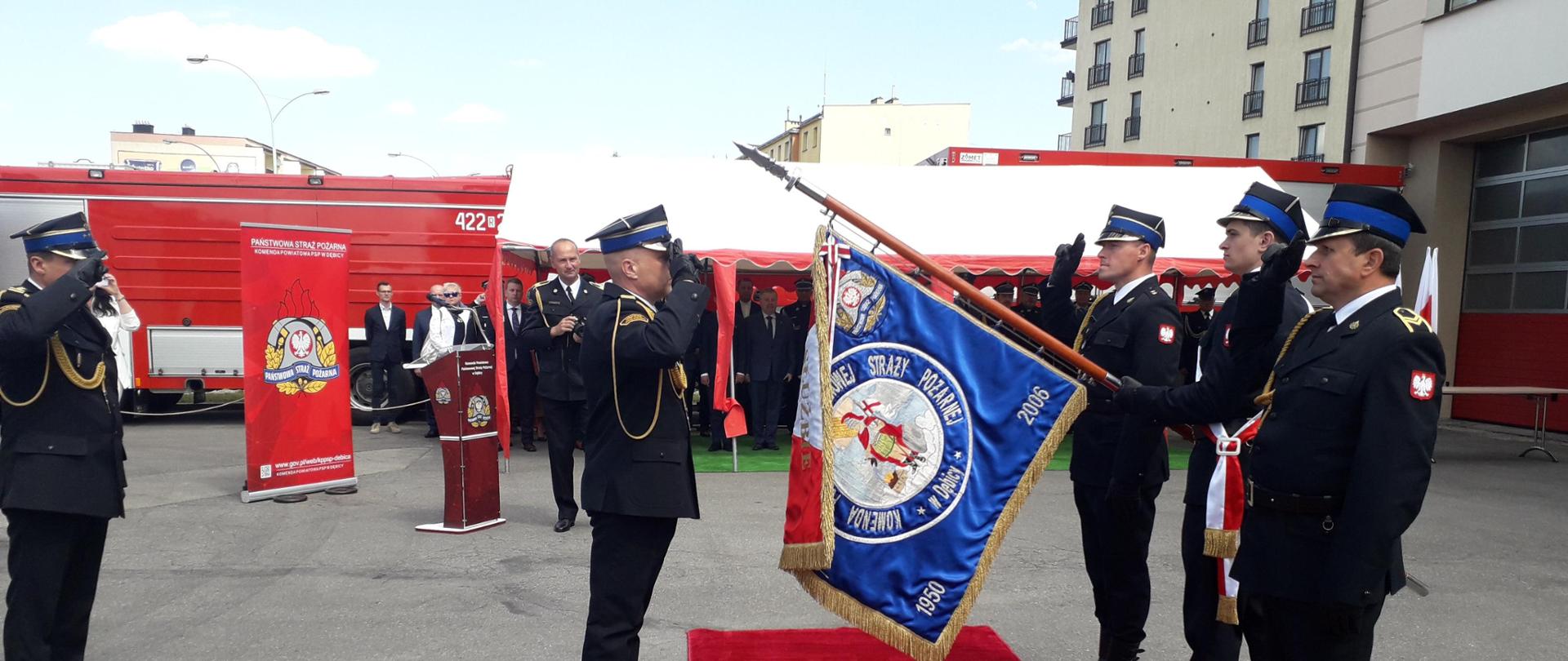 Na zdjęciu komendant powiatowy Państwowej Straży Pożarnej w Dębicy ubrany w mundur wyjściowy stoi przed sztandarem komendy i oddaje honor. Sztandar pochylony w kierunku komendanta trzyma jeden ze strażaków wchodzących w skład pocztu sztandarowego. W poczcie sztandarowym jest trzech strażaków. Tuż za komendantem powiatowym stoi komendant wojewódzki także w mundurze wyjściowym – oddaje honor. W tle zaproszeni goście oraz samochody pożarnicze. 