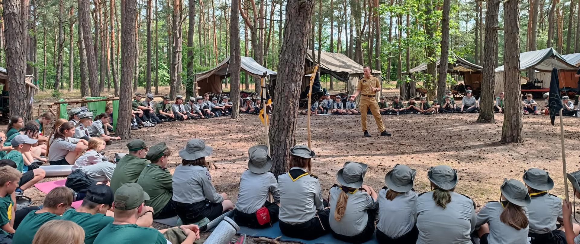 Zdjęcie przedstwia las sosnowy, w którym rozłożony jest obóz harcerski. Na pierwszym planie harcerze siedzą w kręgu, w środku stoi strażak w piaskowym mundurze służbowym. W tle otwarte namioty