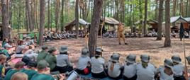 Zdjęcie przedstwia las sosnowy, w którym rozłożony jest obóz harcerski. Na pierwszym planie harcerze siedzą w kręgu, w środku stoi strażak w piaskowym mundurze służbowym. W tle otwarte namioty
